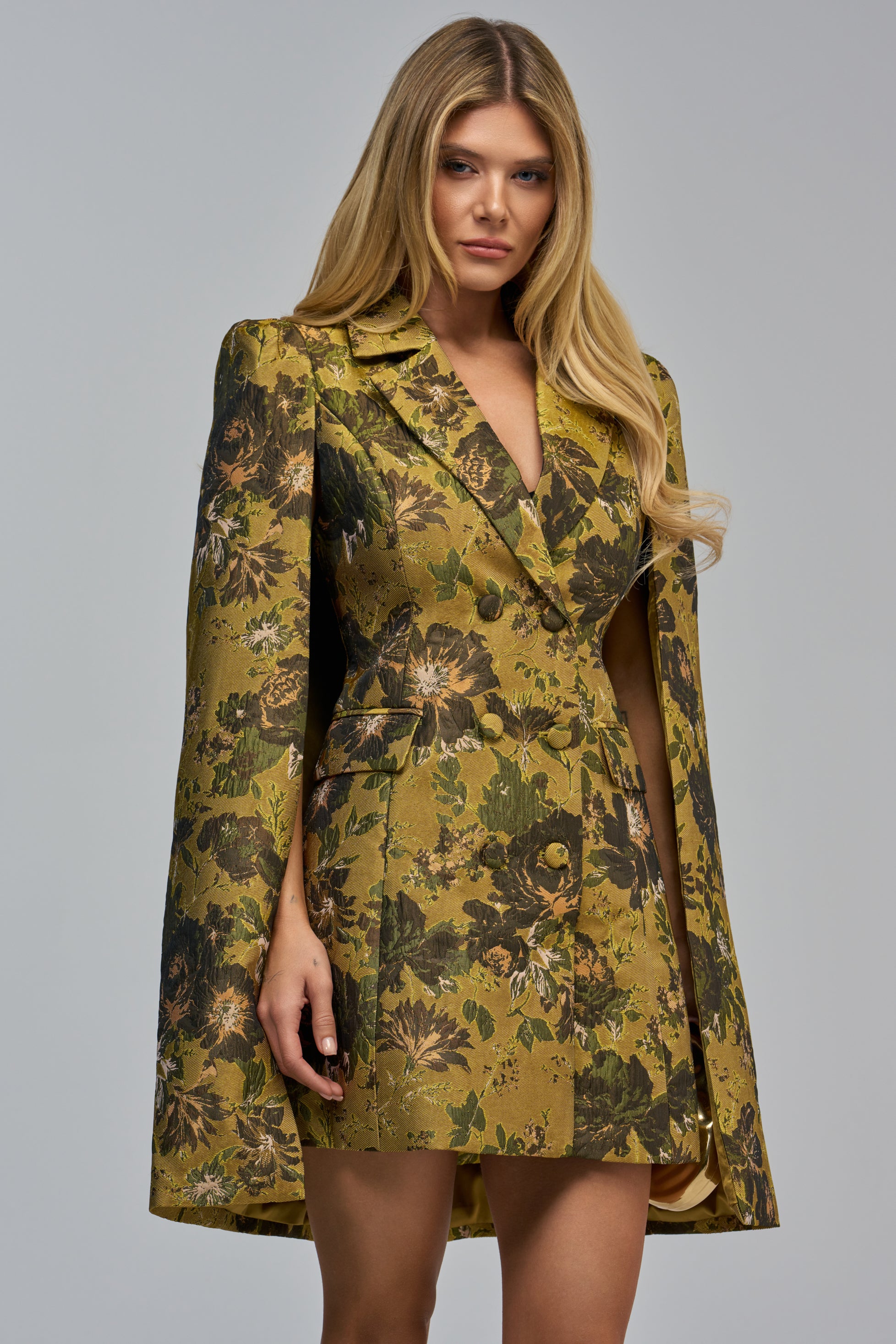 A woman wears the IMANI BROCADE CAPE SLEEVE BLAZER MINI DRESS IN GREEN, an olive green floral jacquard mini dress featuring a double-breasted blazer design with cape sleeves.