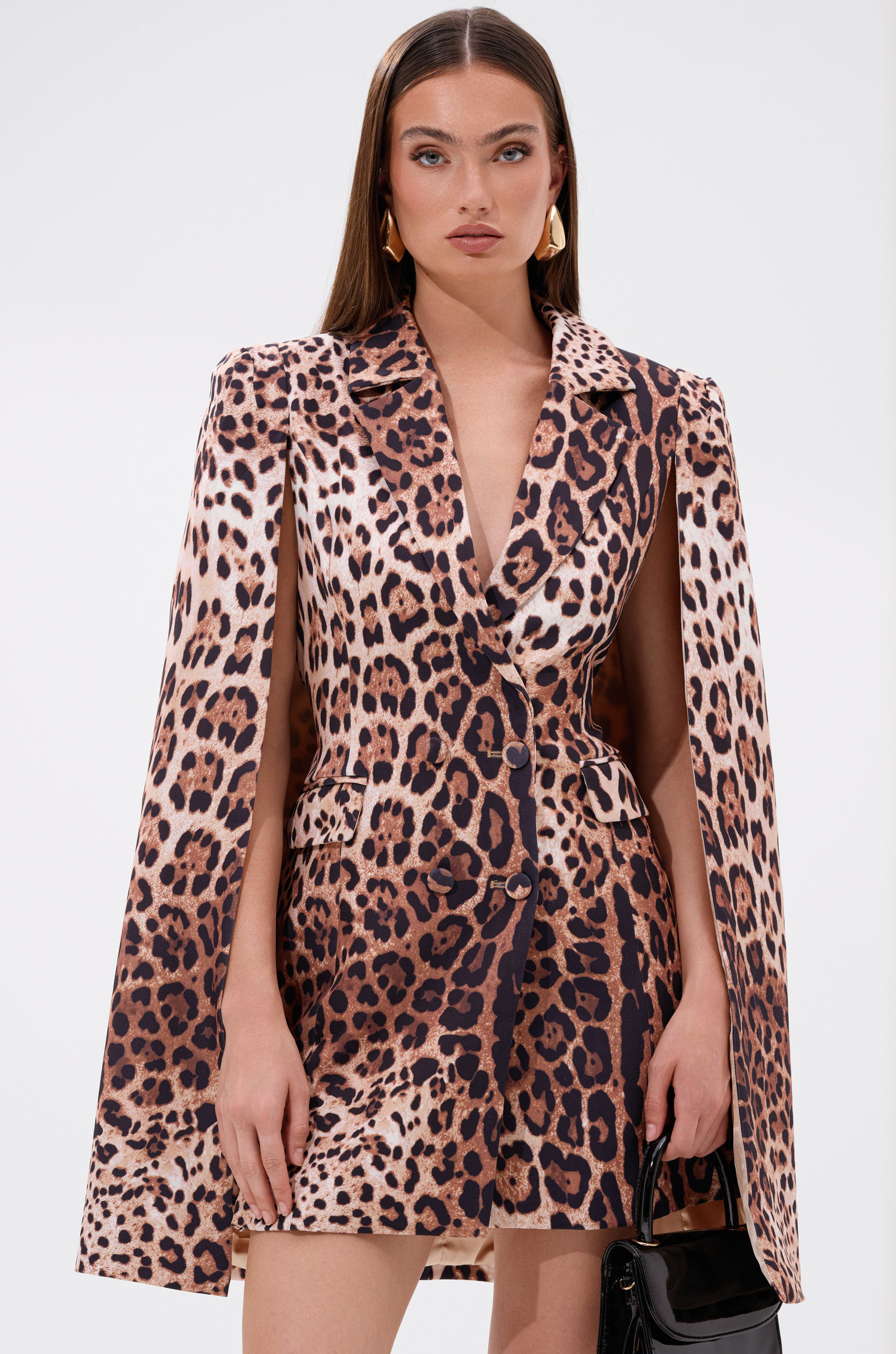 A woman with long straight hair models the AMINAH LEOPARD PRINT BLAZER DRESS with cape sleeves, paired with a small black handbag, posing against a plain white background.