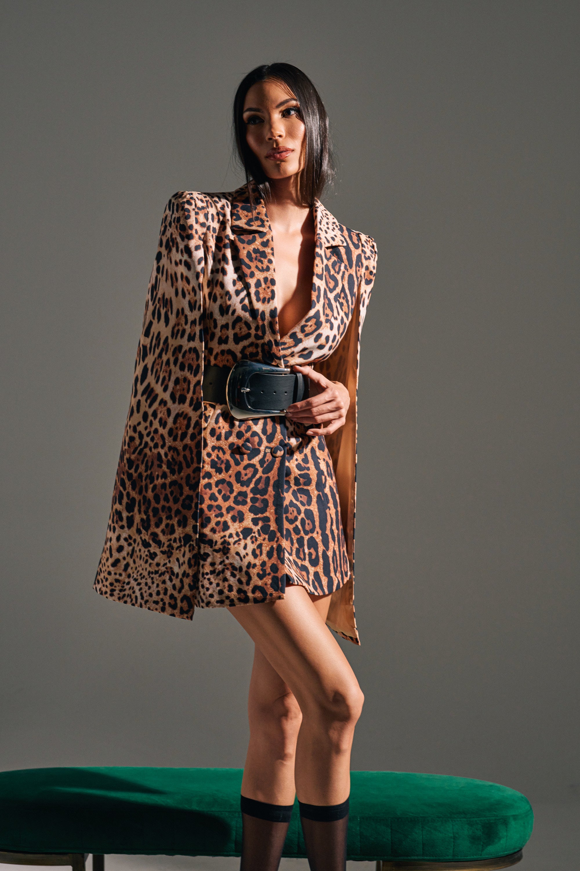 A woman poses confidently in the AMINAH LEOPARD PRINT BLAZER DRESS, featuring dramatic shoulders and a wide black belt, with her hair down and one leg crossed against a gray background and green bench.