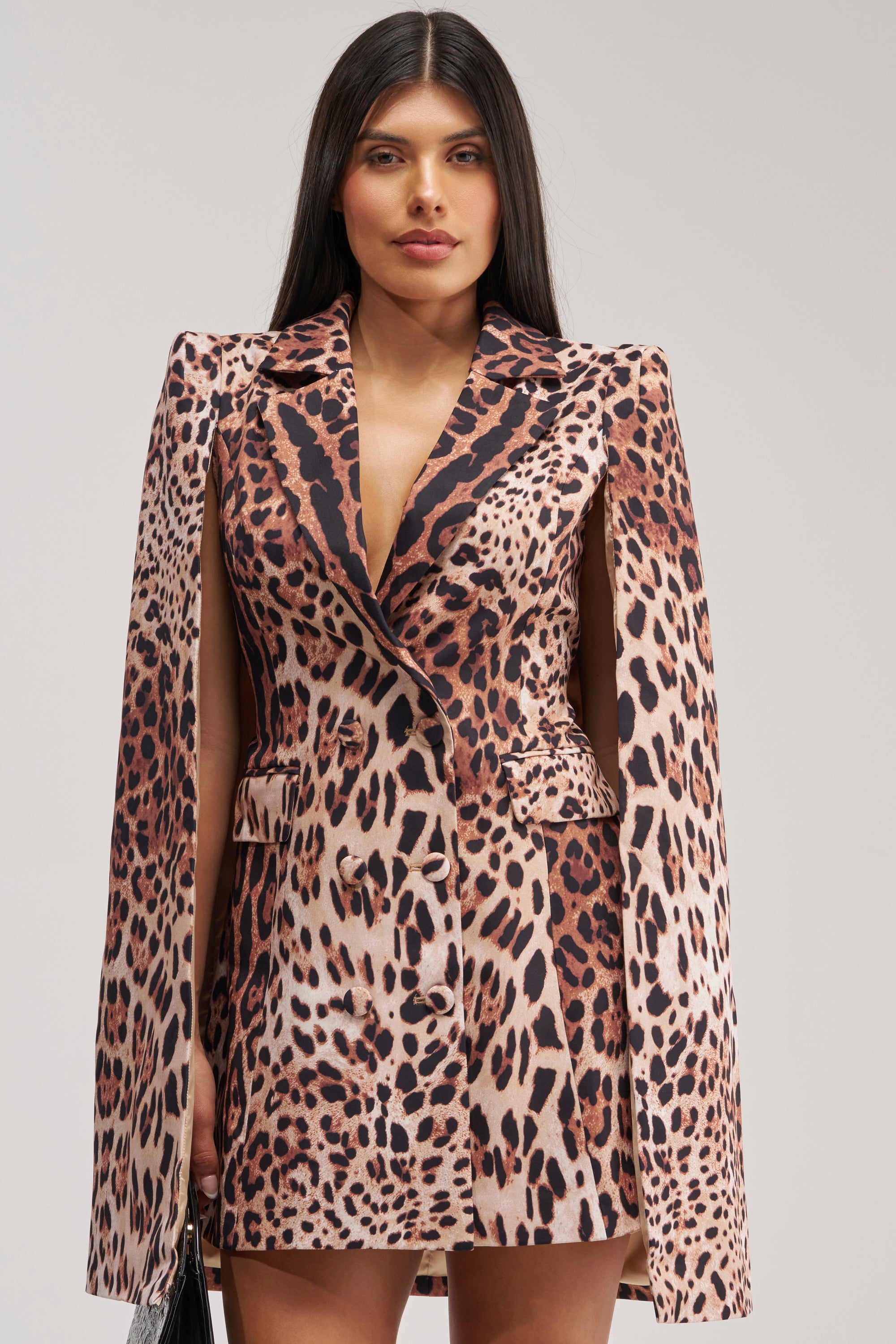 A woman with long dark hair wears the AMINAH LEOPARD PRINT BLAZER DRESS, featuring a cheetah print satin fabric and dramatic cape sleeves, against a plain background. She stands facing forward, holding a small black bag at her side—a striking choice ideal for any cocktail party dress code.