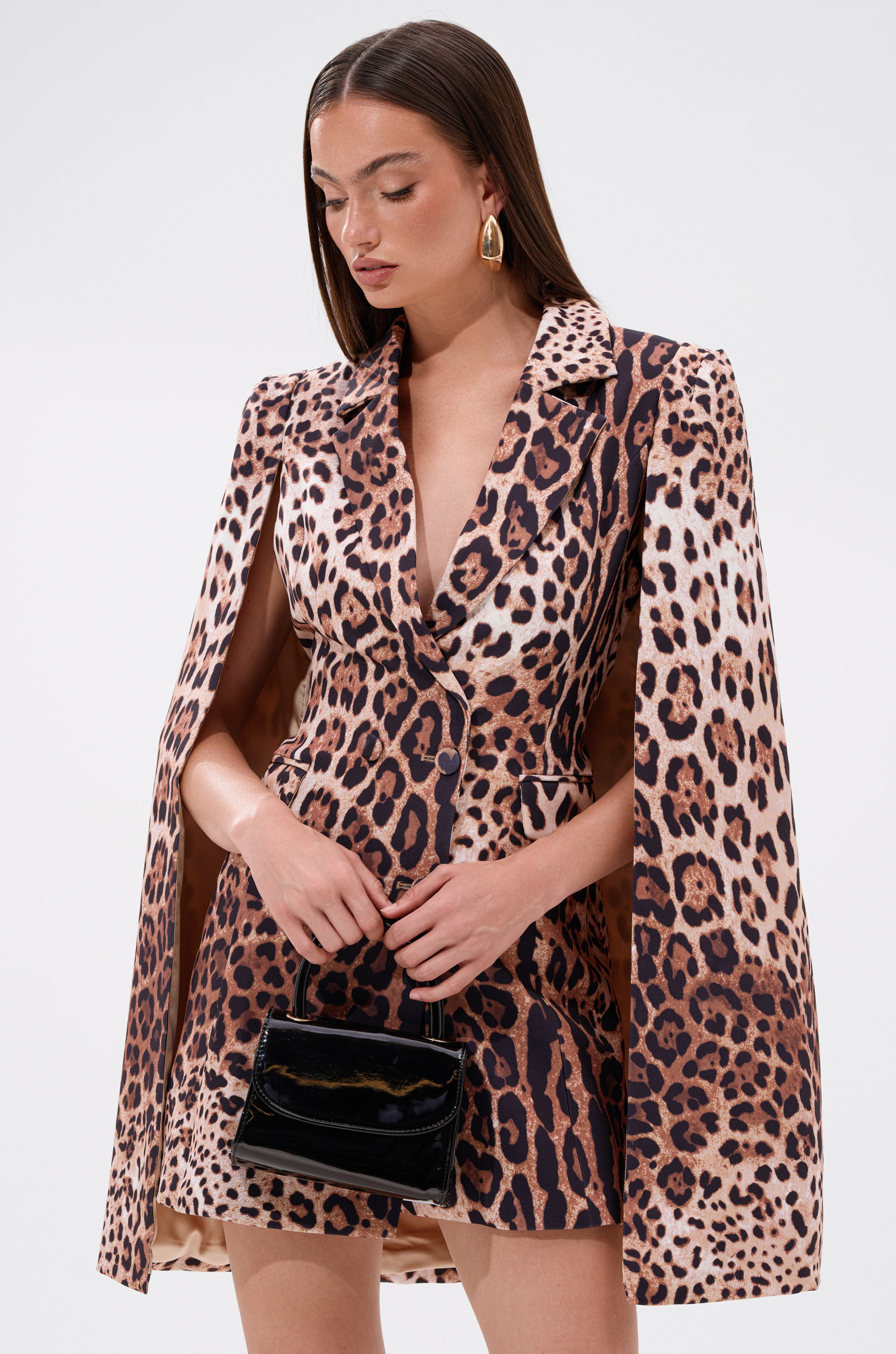 A woman in the AMINAH LEOPARD PRINT BLAZER DRESS with cape sleeves stands against a plain background, holding a small black patent handbag. She has straight brown hair and large gold hoop earrings.