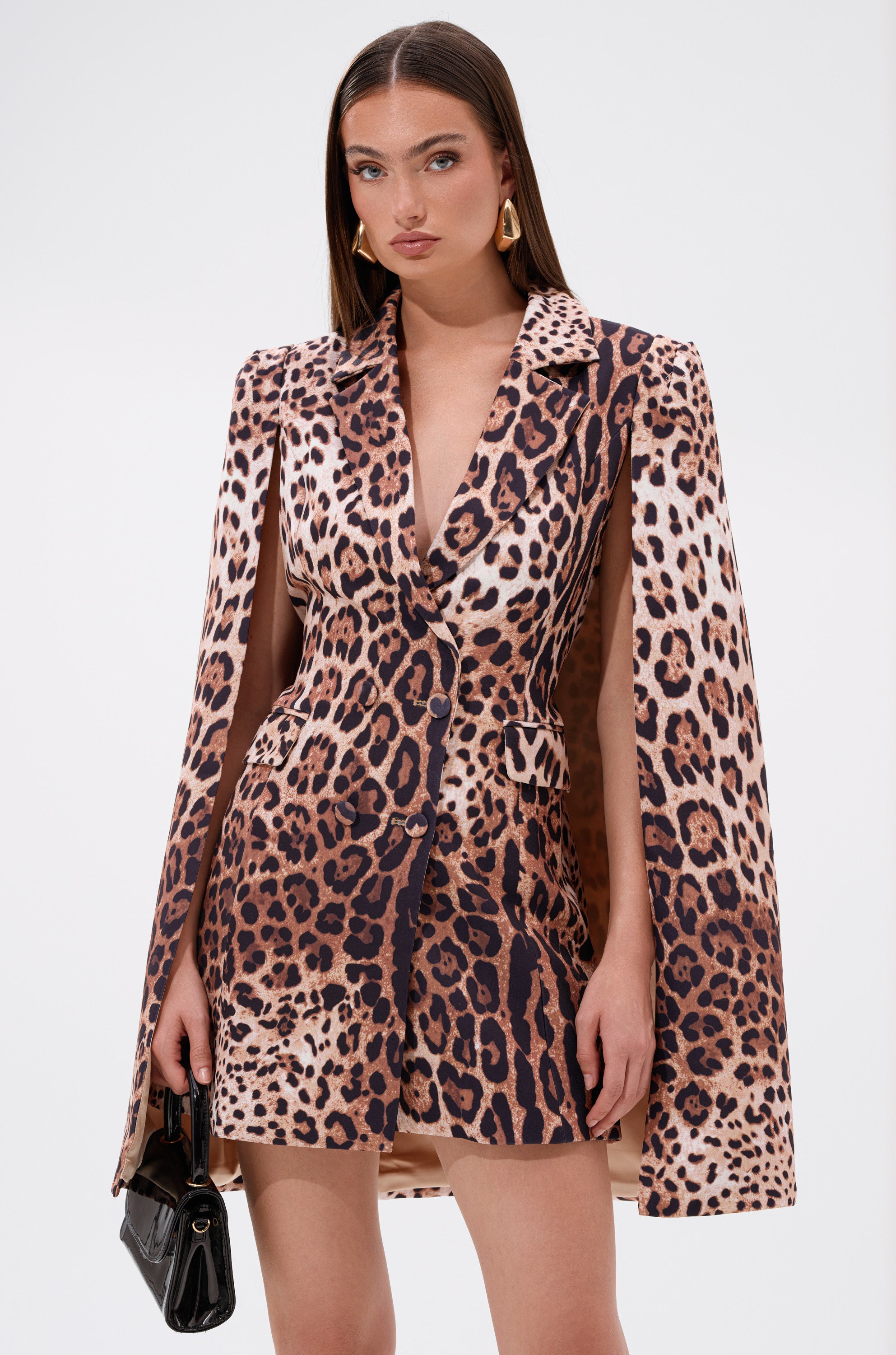 A woman with straight brown hair and gold earrings models the AMINAH LEOPARD PRINT BLAZER DRESS with cape-style sleeves, holding a black handbag against a plain white background.