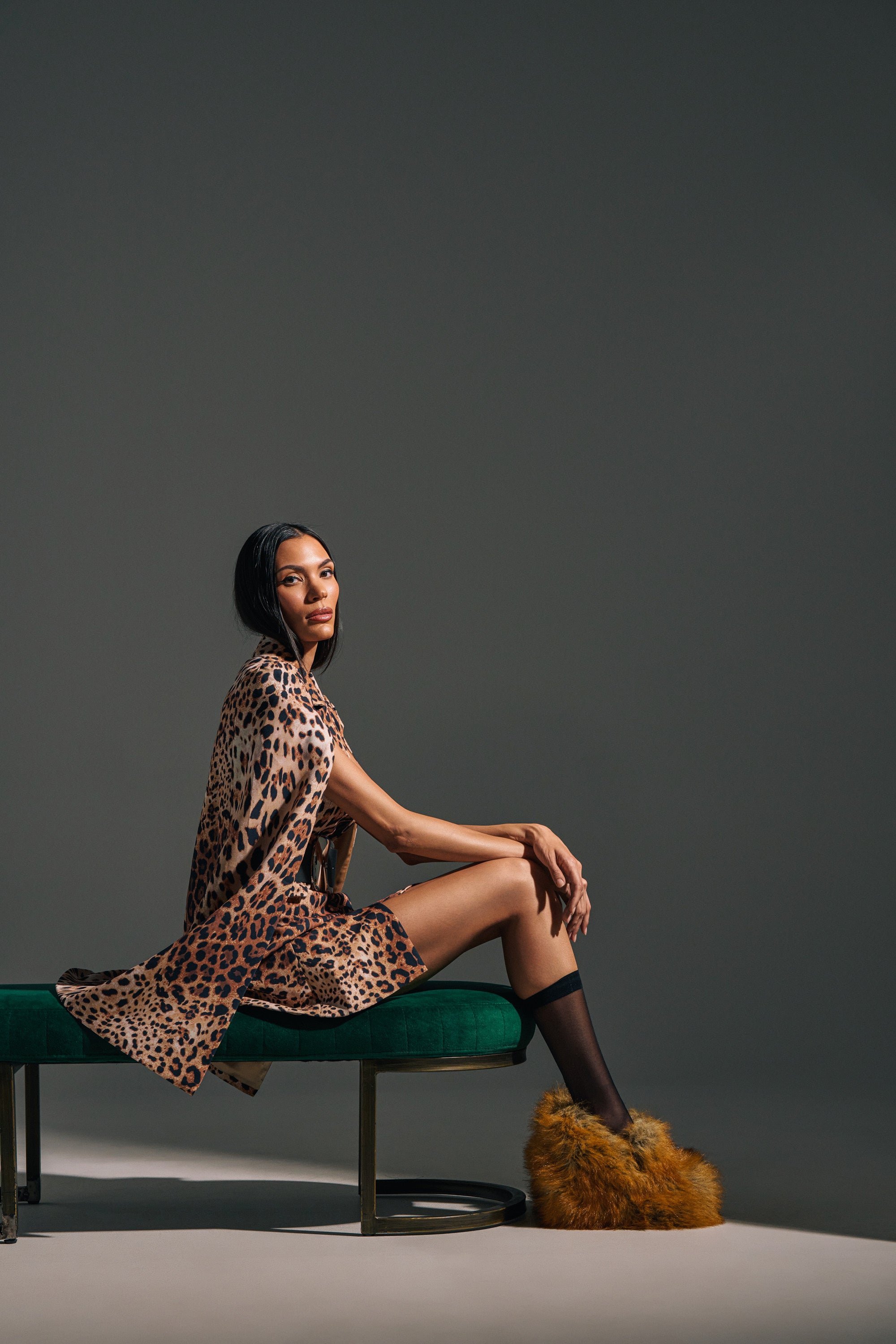 Wearing the AMINAH LEOPARD PRINT BLAZER DRESS, a woman sits sideways on a green bench, gazing at the camera. She pairs the dress with sheer black knee-high socks and fluffy brown slippers against a neutral gray backdrop.