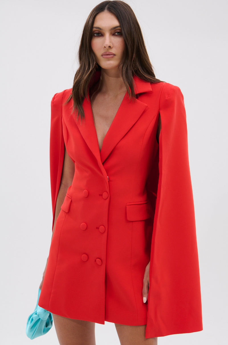 Model wears the A LA MODE MINI BLAZER DRESS IN RED with cape-style sleeves, paired with a light blue clutch, against a plain light gray background.