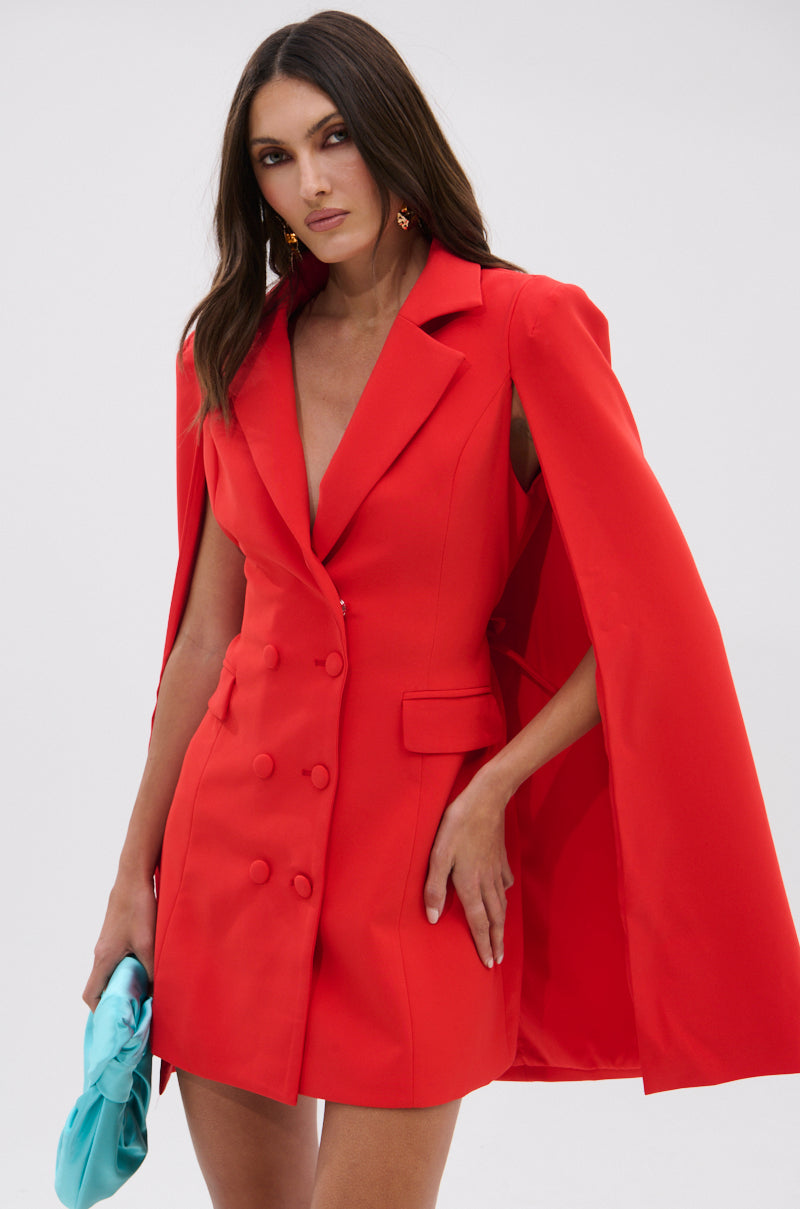 A woman stands confidently against a plain light background, wearing the A LA MODE MINI BLAZER DRESS IN RED with a cape overlay and double-breasted buttons, paired with a light blue clutch.