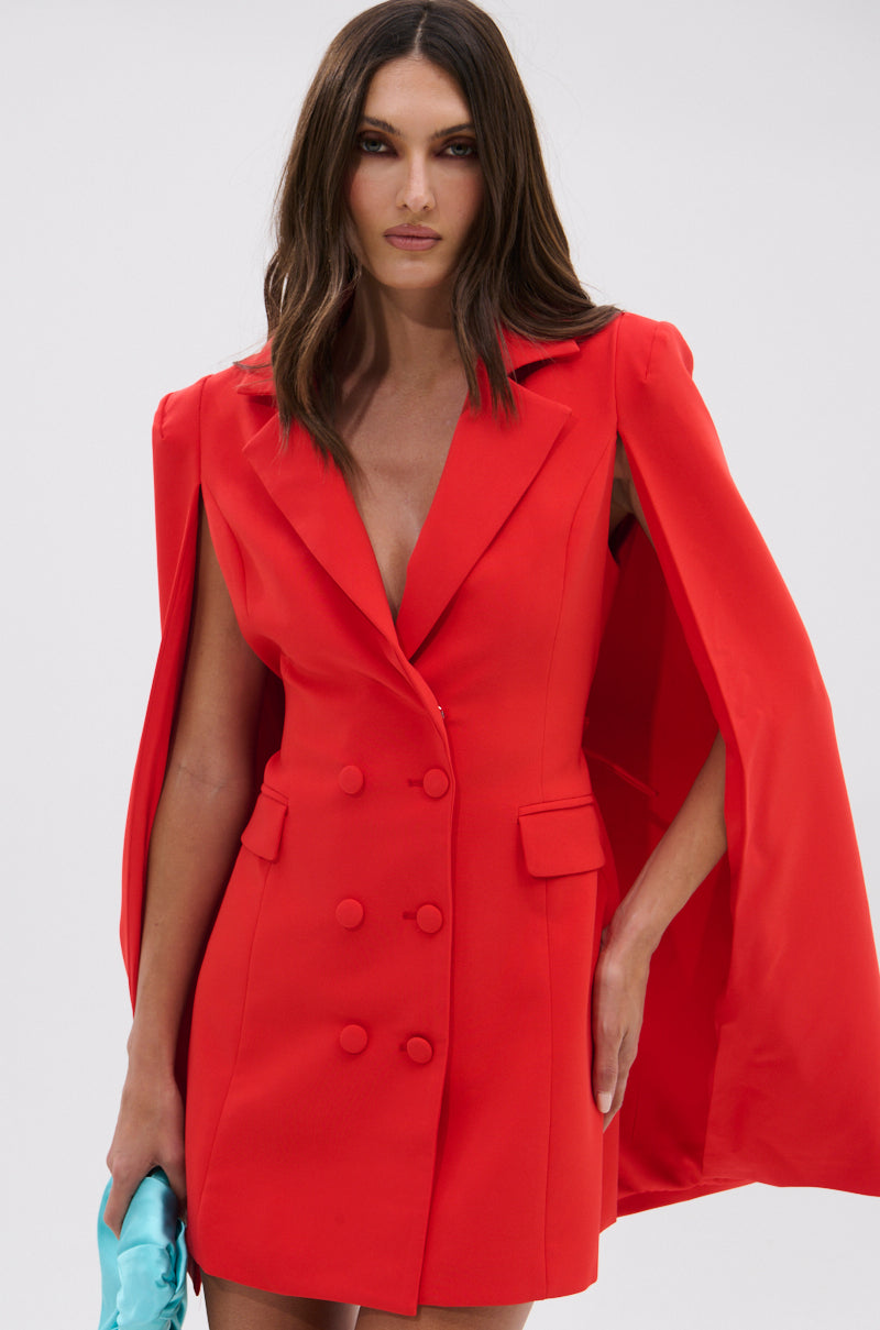 A woman models the A LA MODE MINI BLAZER DRESS IN RED, featuring a chic cape overlay, as she stands against a plain background holding a light blue clutch in her left hand.