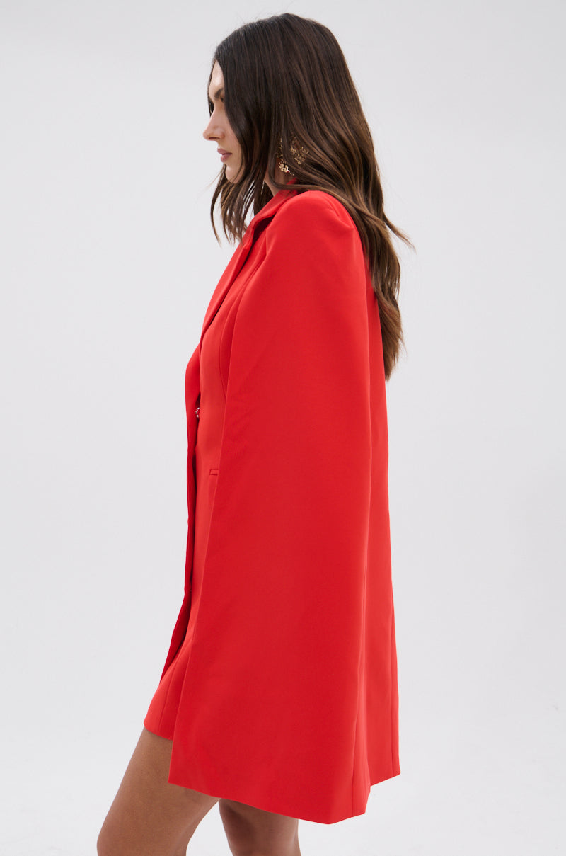 A woman with long brown hair stands in profile against a light gray background, modeling the A LA MODE MINI BLAZER DRESS IN RED featuring a cape overlay and matching accents.