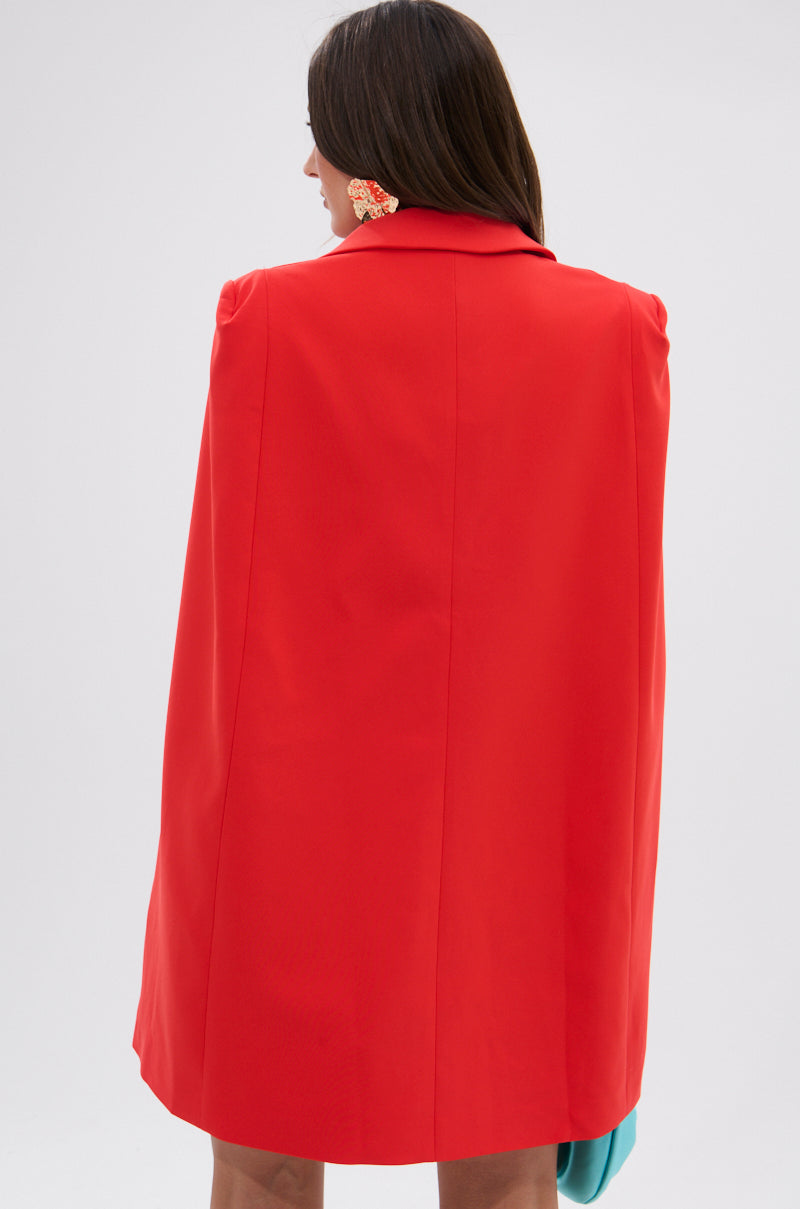 Seen from behind, a woman with long brown hair wears the A LA MODE MINI BLAZER DRESS IN RED with a cape overlay, a flower-shaped earring, and holds a turquoise item in her left hand against a plain light background.