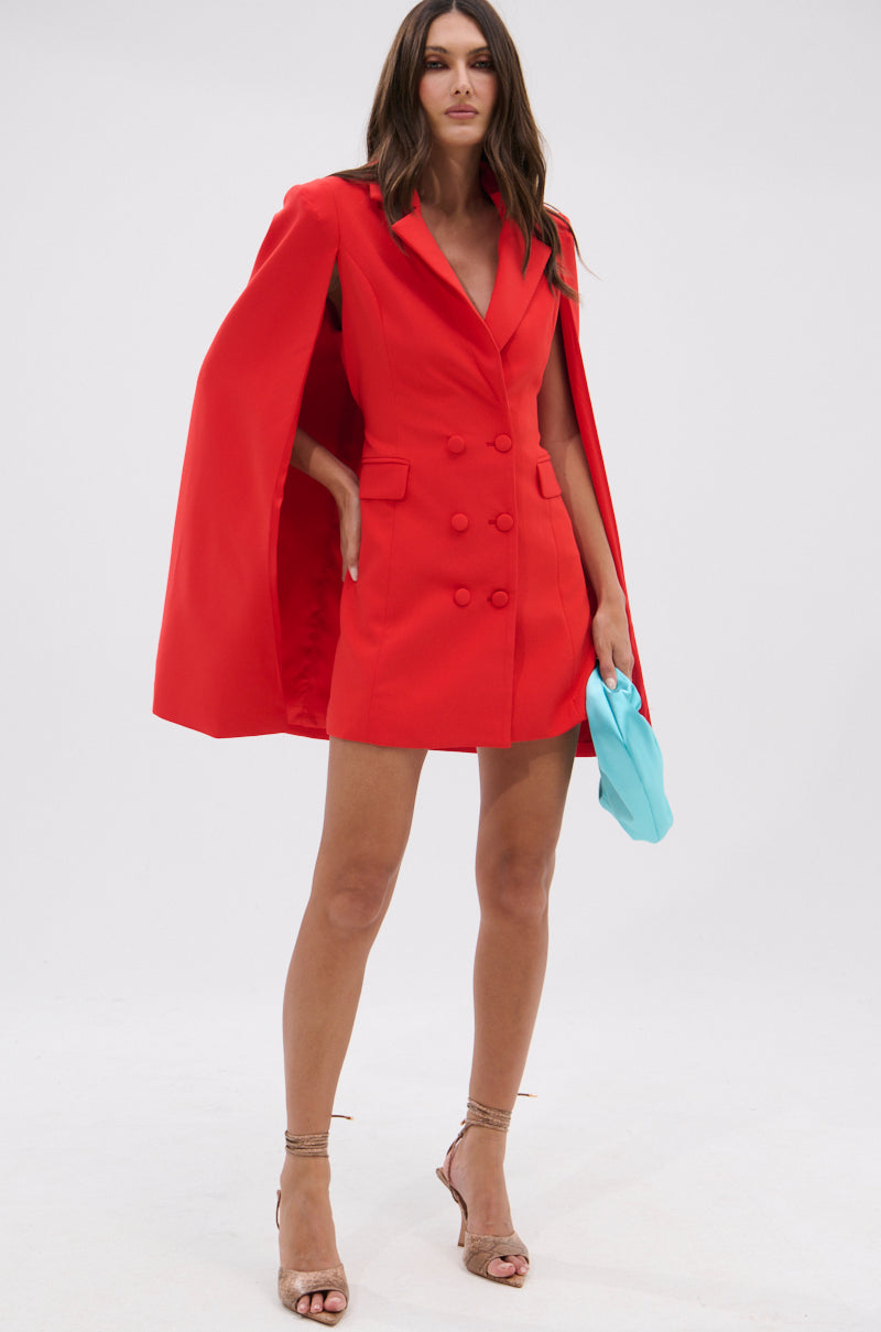 A woman stands confidently in the A LA MODE MINI BLAZER DRESS IN RED, featuring a bold cape overlay. She accessorizes with a light blue clutch and strappy high heels, set against a simple white background.