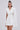 A woman with long brown hair in a high bun wears the A LA MODE MINI BLAZER DRESS IN WHITE with cape sleeves, standing against a plain light background and looking confidently at the camera.