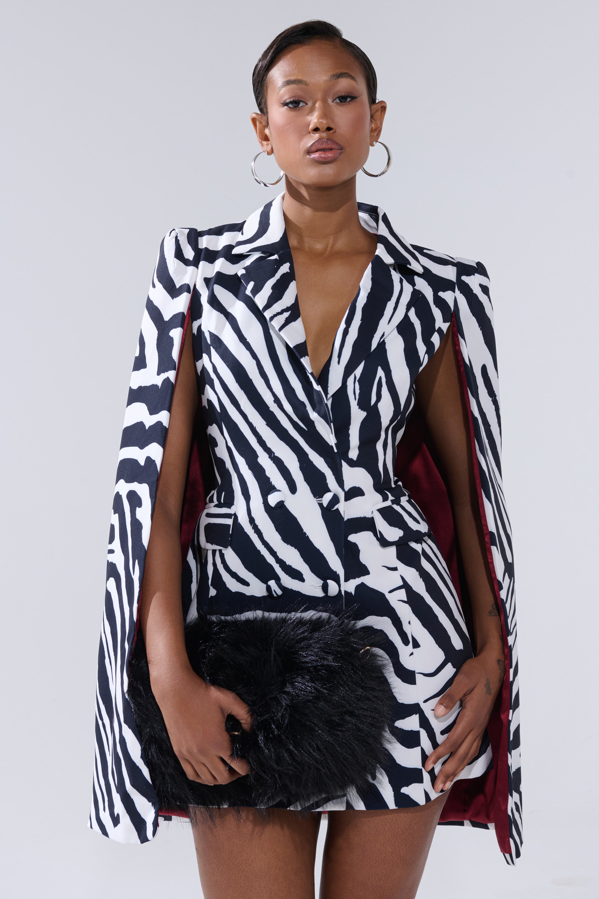A woman confidently poses against a light gray background in the WILD SIDE PRINTED BLAZER DRESS, a chic black and white zebra print mini dress with cape sleeves, paired with a black furry clutch for an unforgettable statement look.