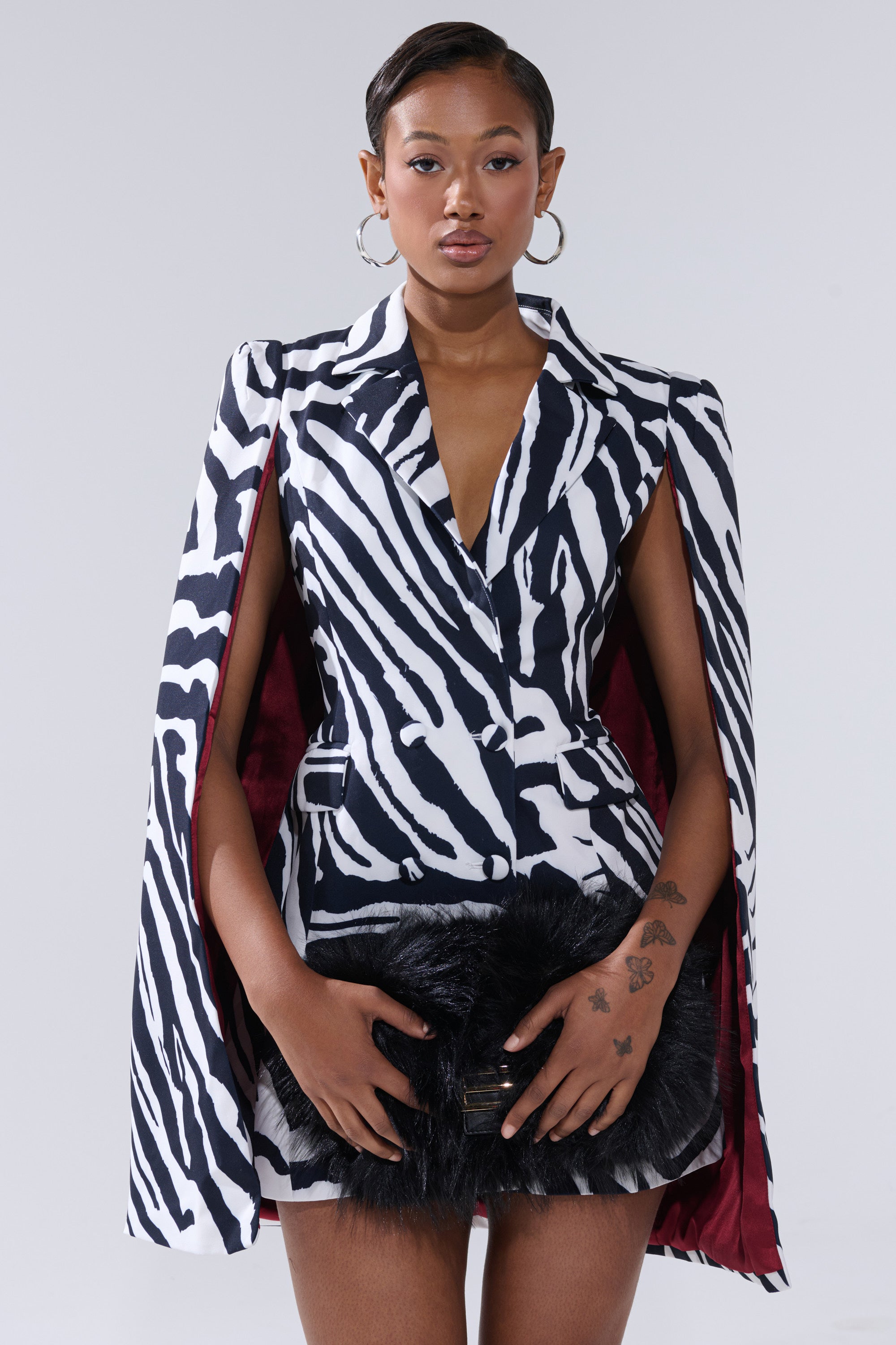 A woman poses against a plain backdrop in the WILD SIDE PRINTED BLAZER DRESS, featuring a bold zebra pattern and cape sleeves. She accessorizes with a black fur clutch, hoop earrings, sleek short hair, and hand tattoos.