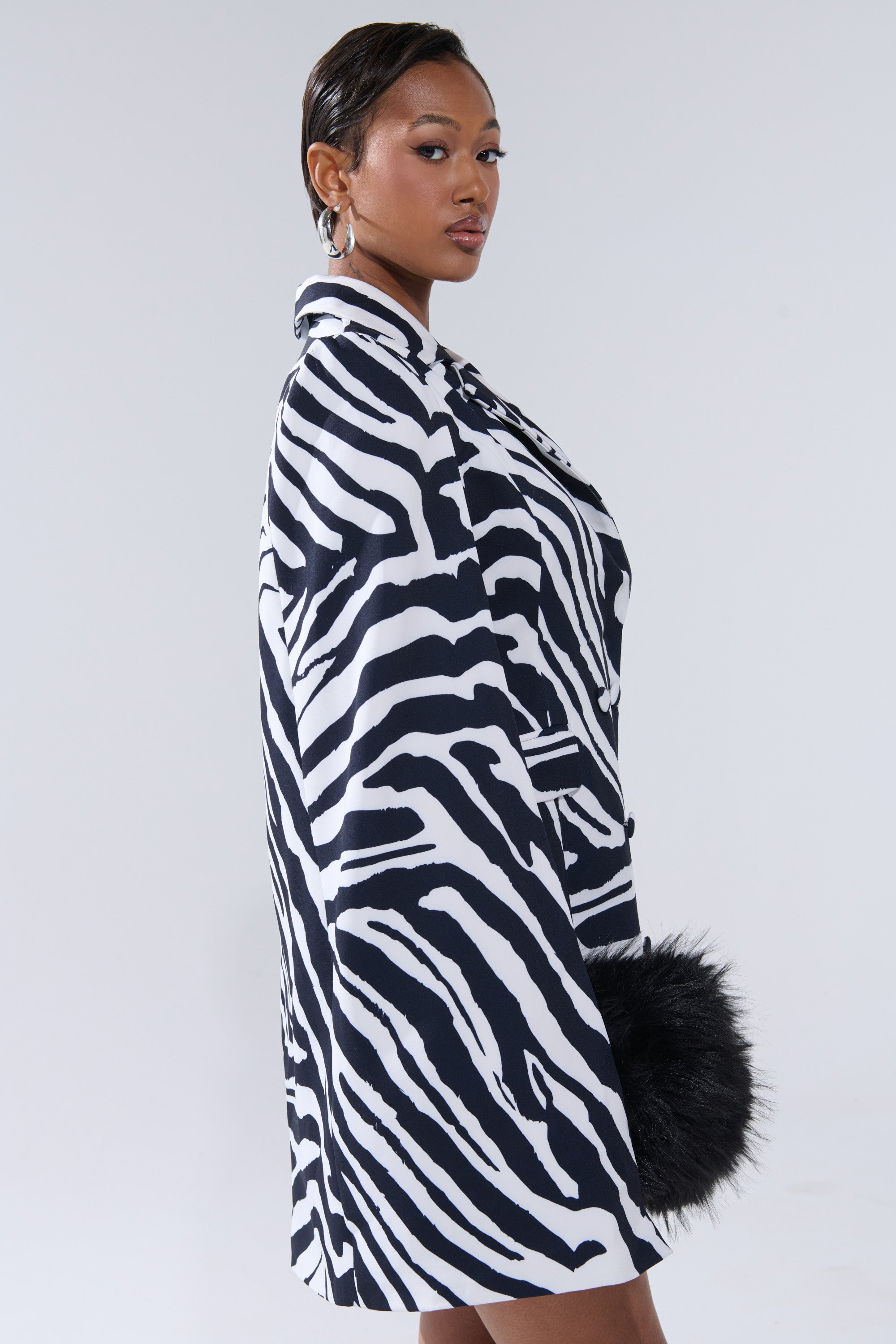 A woman models the WILD SIDE PRINTED BLAZER DRESS with a black furry clutch, short slicked-back hair, and large hoop earrings, posed against a plain light background.