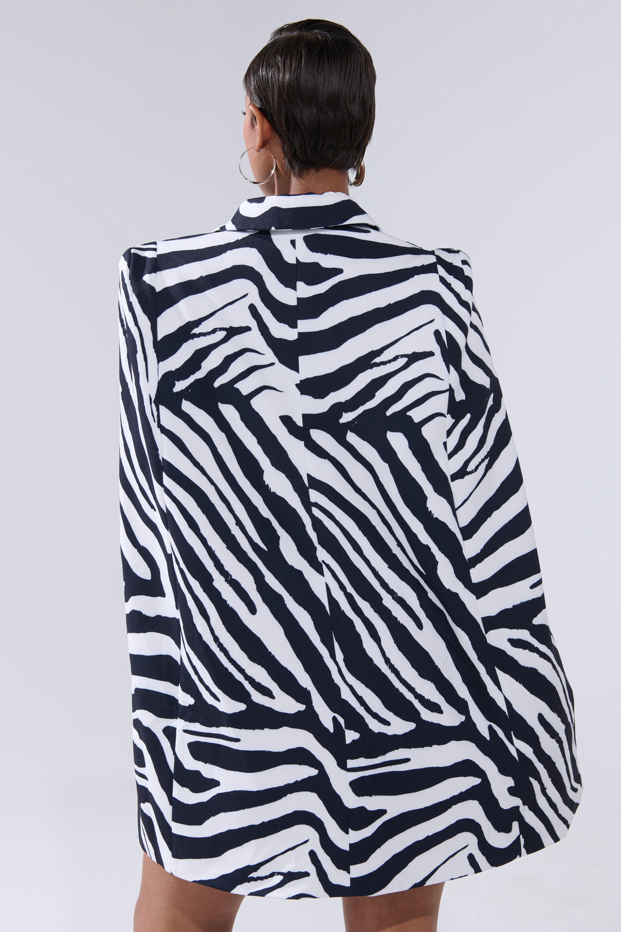 A person with short hair, seen from behind, models the WILD SIDE PRINTED BLAZER DRESS featuring a black and white zebra print. The background is plain light gray.