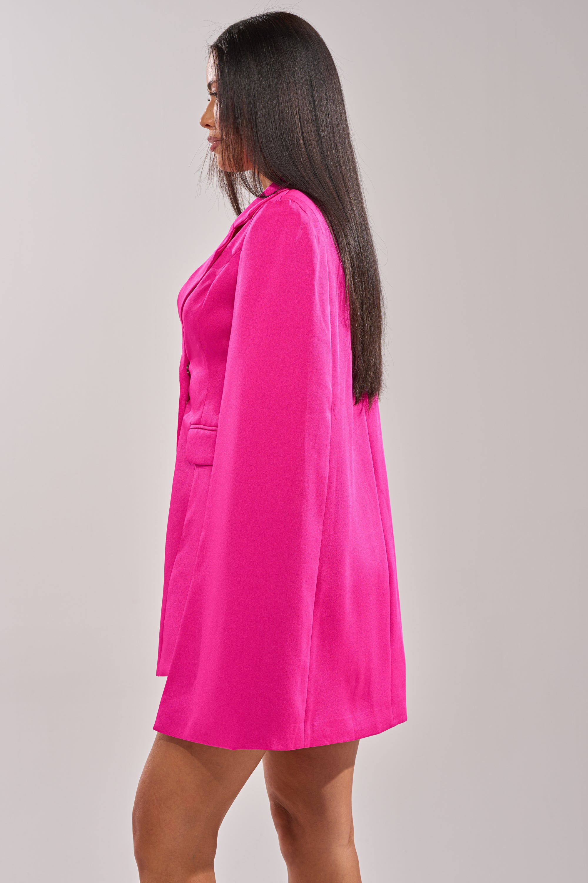 A woman in profile wears the A LA MODE MINI BLAZER DRESS IN PINK, a structured mini cape blazer dress, with long straight hair and a neutral background. 3