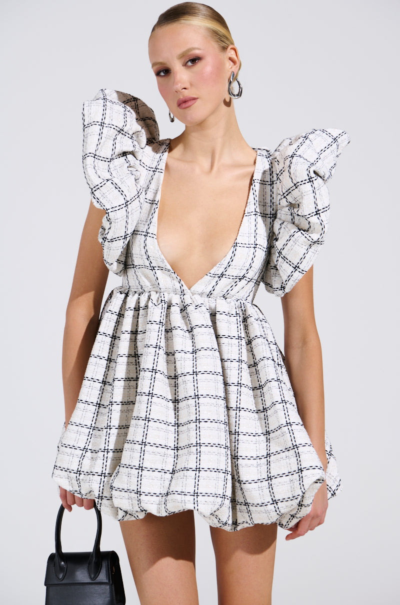 A person poses in the chic THAT GIRL TWEED MINI DRESS with ruffled sleeves and a deep V-neck. Their slicked-back hairstyle, hoop earrings, and small black handbag create a stylish look against the light-colored background.