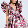 A woman poses confidently in the MAIN CHARACTER ENERGY BROCADE MINI DRESS IN SILVER, featuring a short, floral-patterned design with voluminous ruffled sleeves, shades of pink, purple, and gray, and a deep plunging neckline. Her long brown hair flows against a light background.