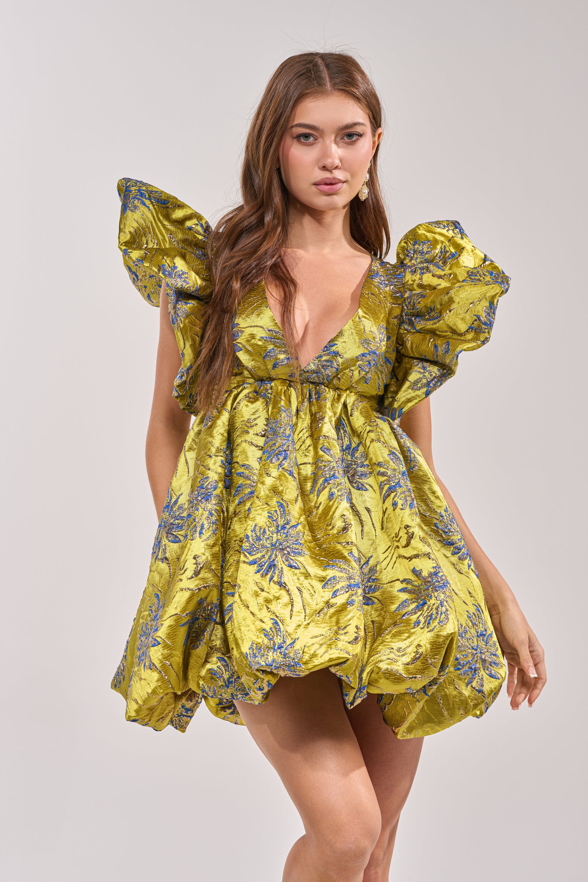 A woman wears the MAIN CHARACTER ENERGY BROCADE MINI DRESS IN GREEN, featuring dramatic ruffled sleeves and a bold floral pattern—an eye-catching cocktail party statement against a plain light background.
