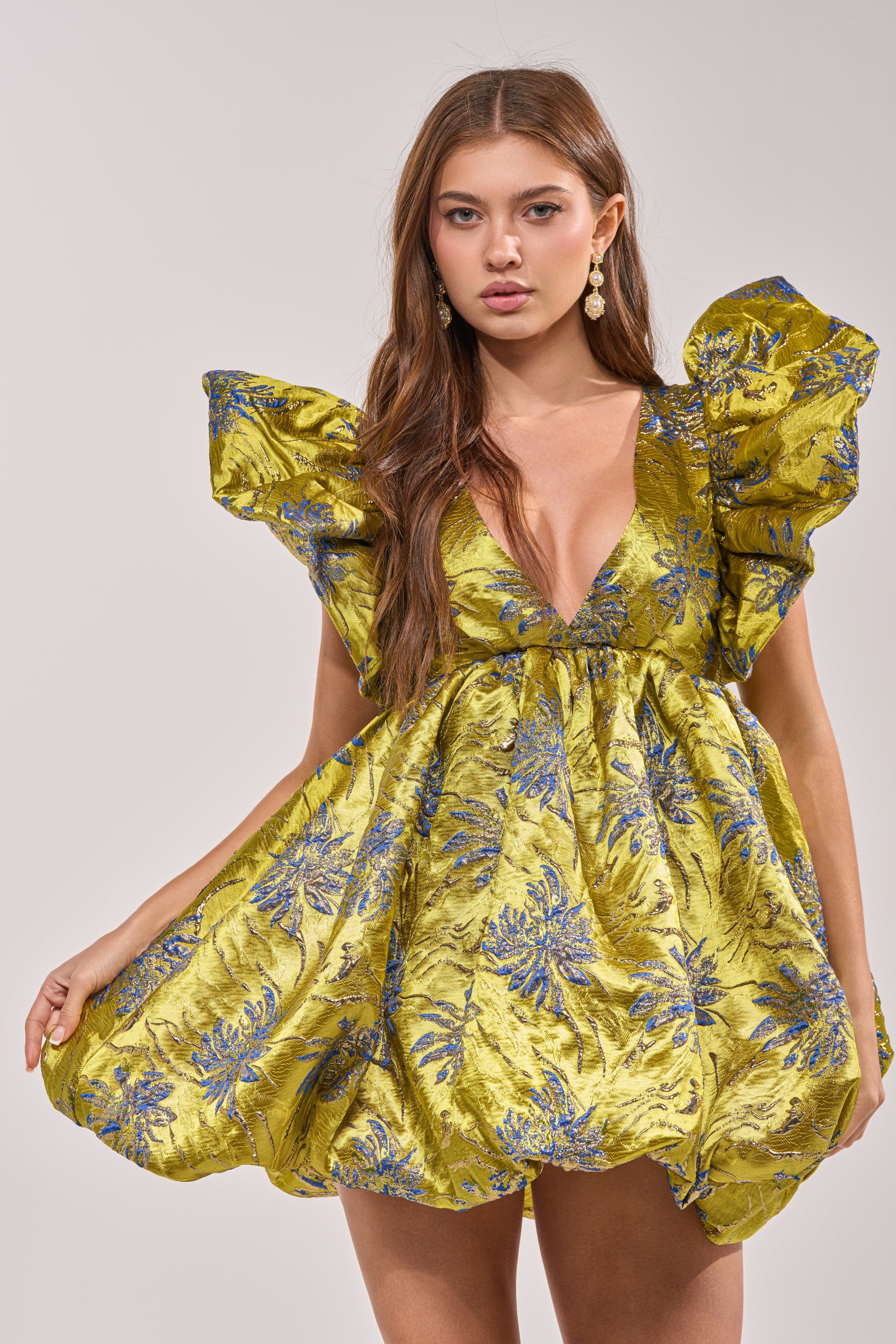 A woman wearing the MAIN CHARACTER ENERGY BROCADE MINI DRESS IN GREEN, a shiny, short mini dress with ruffled sleeves, stands against a plain background and looks directly at the camera.