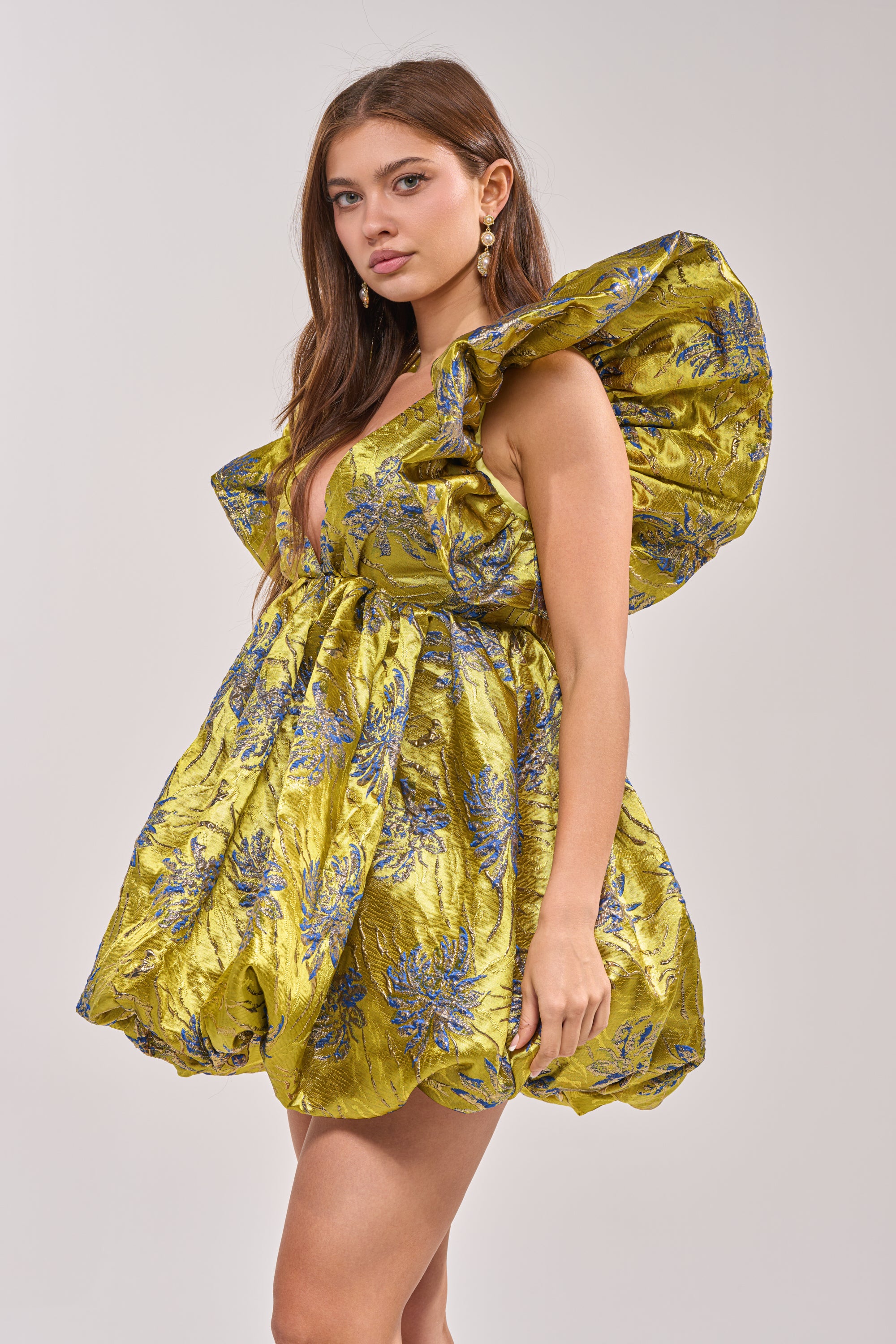 A woman poses in the MAIN CHARACTER ENERGY BROCADE MINI DRESS IN GREEN, featuring blue floral patterns, a deep V-neckline, ruffled shoulders, and statement sleeves. She has long brown hair and wears dangling earrings.