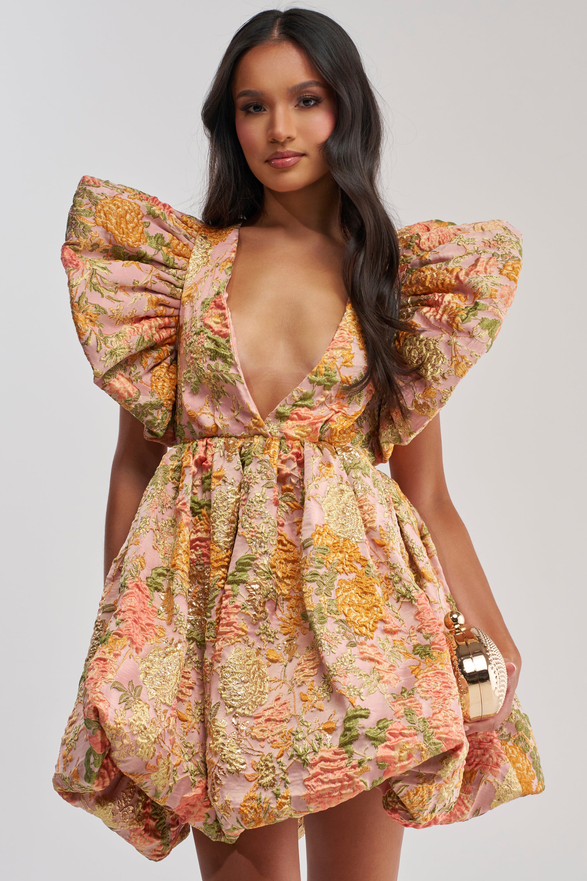 A woman with long, wavy dark hair wears the MAIN CHARACTER ENERGY BROCADE MINI DRESS IN PEACH—a floral mini with dramatic puffed sleeves, a deep V-neckline, and pink, gold, and green tones. She carries a small handbag.