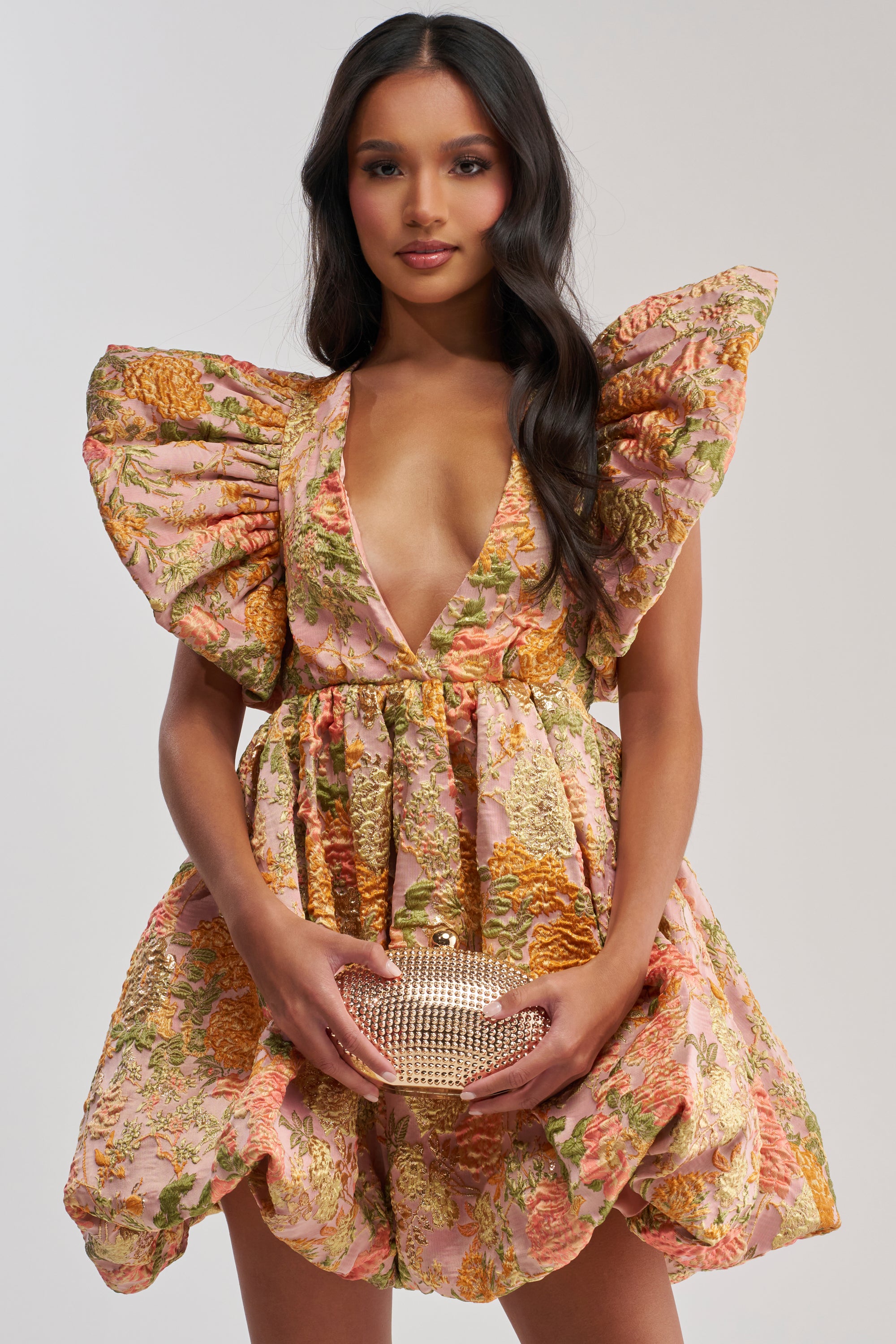 A woman in the MAIN CHARACTER ENERGY BROCADE MINI DRESS IN PEACH, featuring dramatic puffed sleeves and a deep V-neckline, holds a sparkly clutch bag against a plain background.