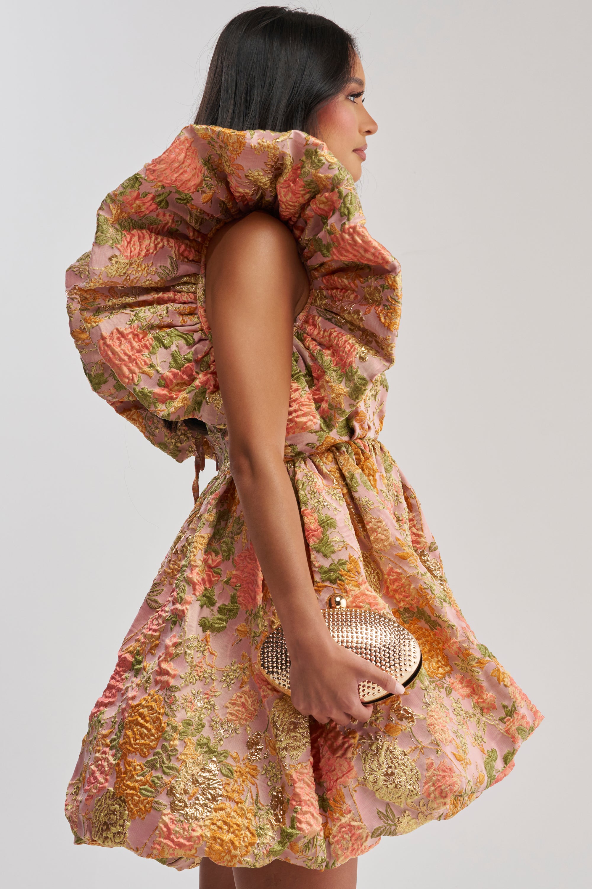 A woman in the MAIN CHARACTER ENERGY BROCADE MINI DRESS IN PEACH, featuring pink, gold, and green florals with a large ruffled collar, stands in profile holding a gold clutch against a plain light background.