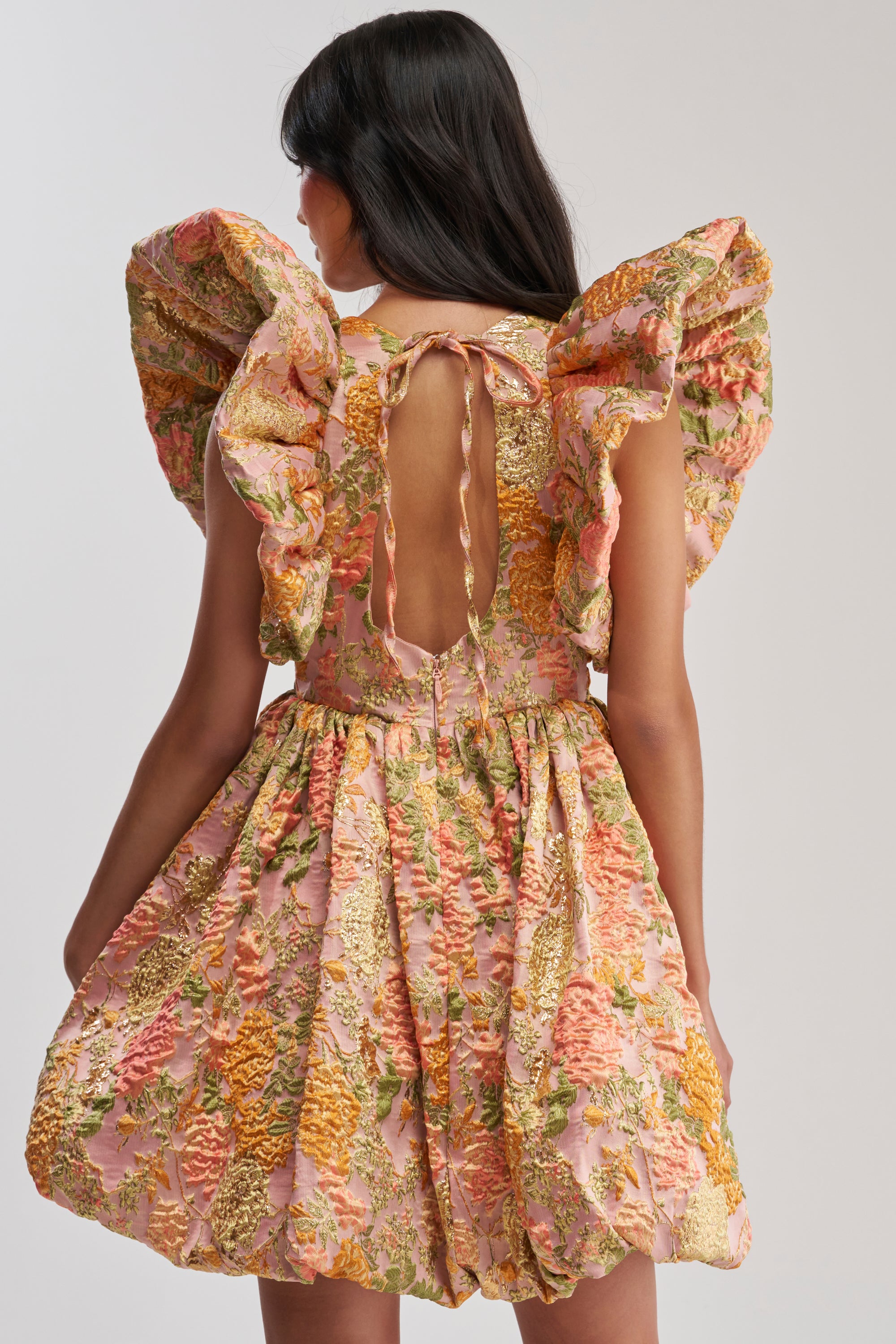 A woman faces away in the MAIN CHARACTER ENERGY BROCADE MINI DRESS IN PEACH, featuring a voluminous skirt with ornate embroidery, large ruffled sleeves, and an open back tied with a ribbon—ideal for cocktail parties.