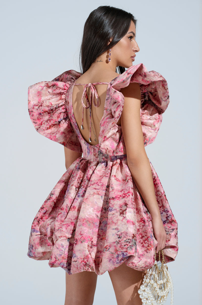 The woman, wearing the BELLE OF THE BALL PRINTED MINI DRESS featuring a pink floral brocade print and a deep neckline, holds a beaded handbag. Her long dark hair flows down her back against a soft blue background. 4
