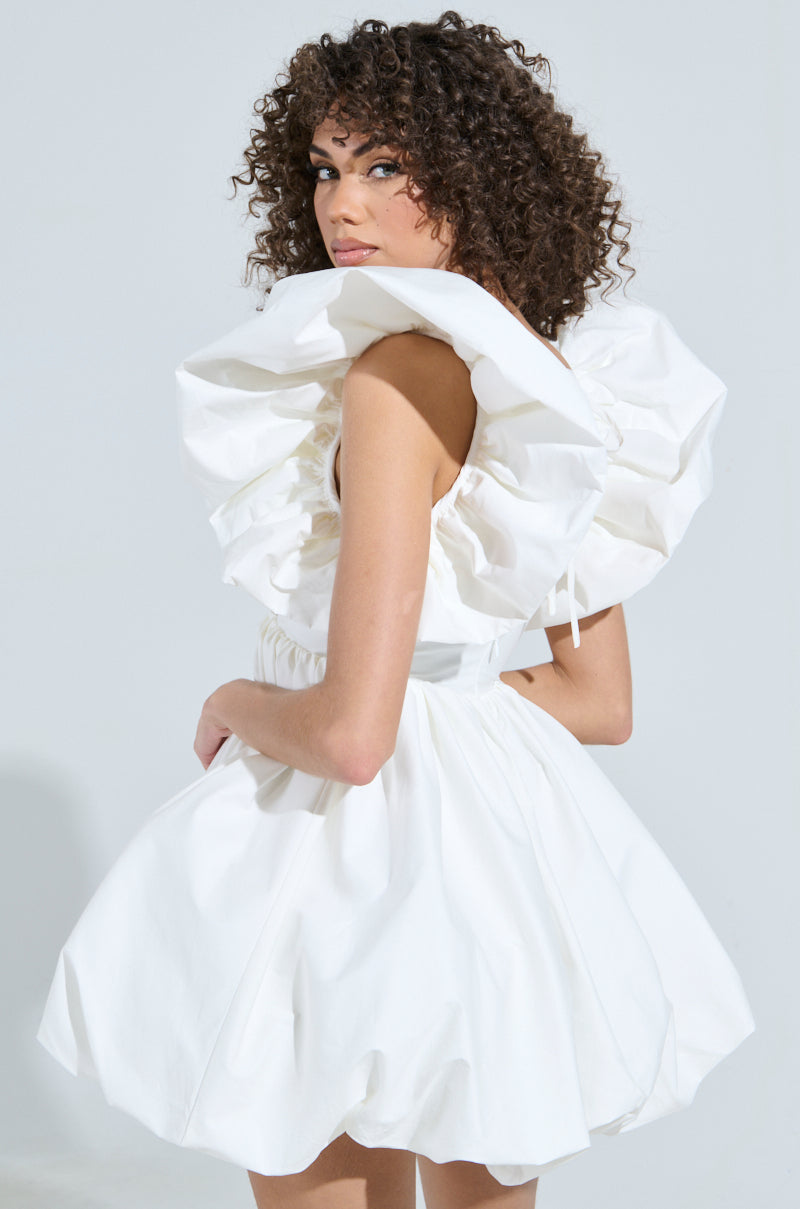 A woman with curly hair exudes main character energy in the MAIN CHARACTER ENERGY MINI DRESS IN WHITE, featuring dramatic ruffles around the shoulders and a flared skater silhouette. She's looking over her shoulder against a simple light gray backdrop. 0