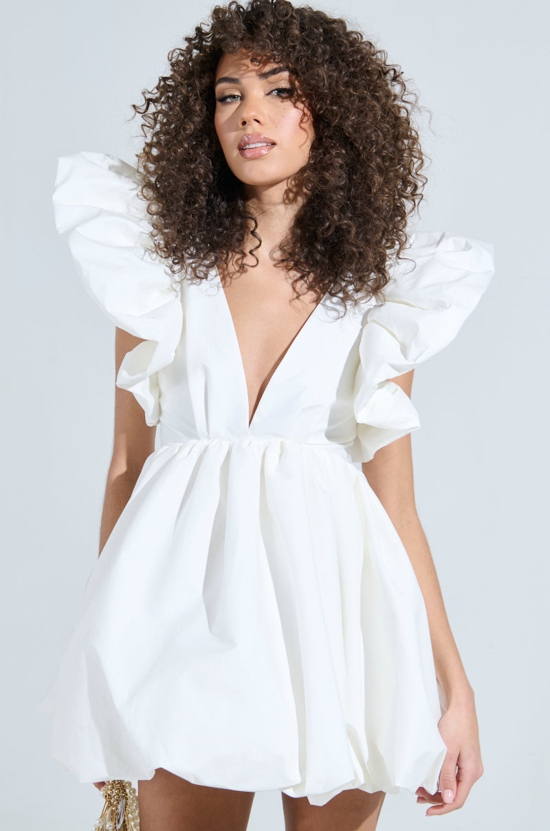 A person with curly hair exudes main character energy in the MAIN CHARACTER ENERGY MINI DRESS IN WHITE with voluminous, ruffled sleeves and a deep V-neckline. They confidently stand against a plain background, gazing directly at the camera. 1