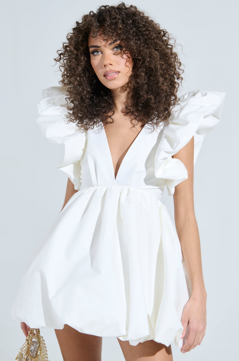 A woman with curly hair exudes main character energy in the MAIN CHARACTER ENERGY MINI DRESS IN WHITE with large ruffled sleeves and a deep V-neckline. She confidently holds a small gold purse against a light-colored background. 2