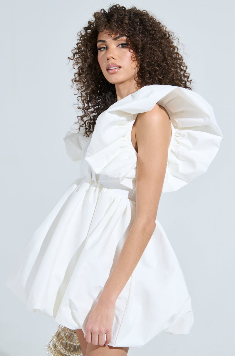 Curly-haired person wears the MAIN CHARACTER ENERGY MINI DRESS IN WHITE, showcasing oversized ruffled sleeves and a flared skater silhouette against a plain background, emphasizing its dramatic design and standout style. 3