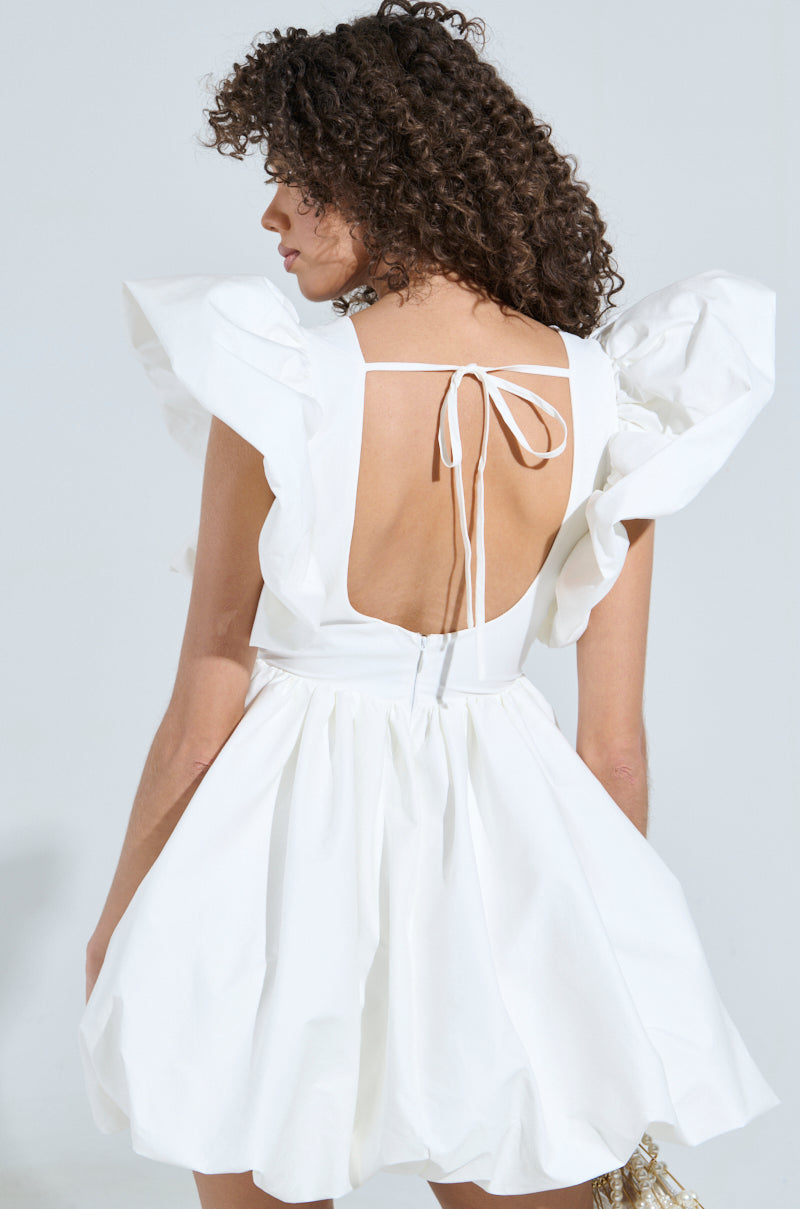 With curly hair, a person exudes main character energy in the MAIN CHARACTER ENERGY MINI DRESS IN WHITE. It showcases large ruffled sleeves, an open back with tie detailing, and a skater silhouette. They stand against a light gray background, facing away from the camera. 4