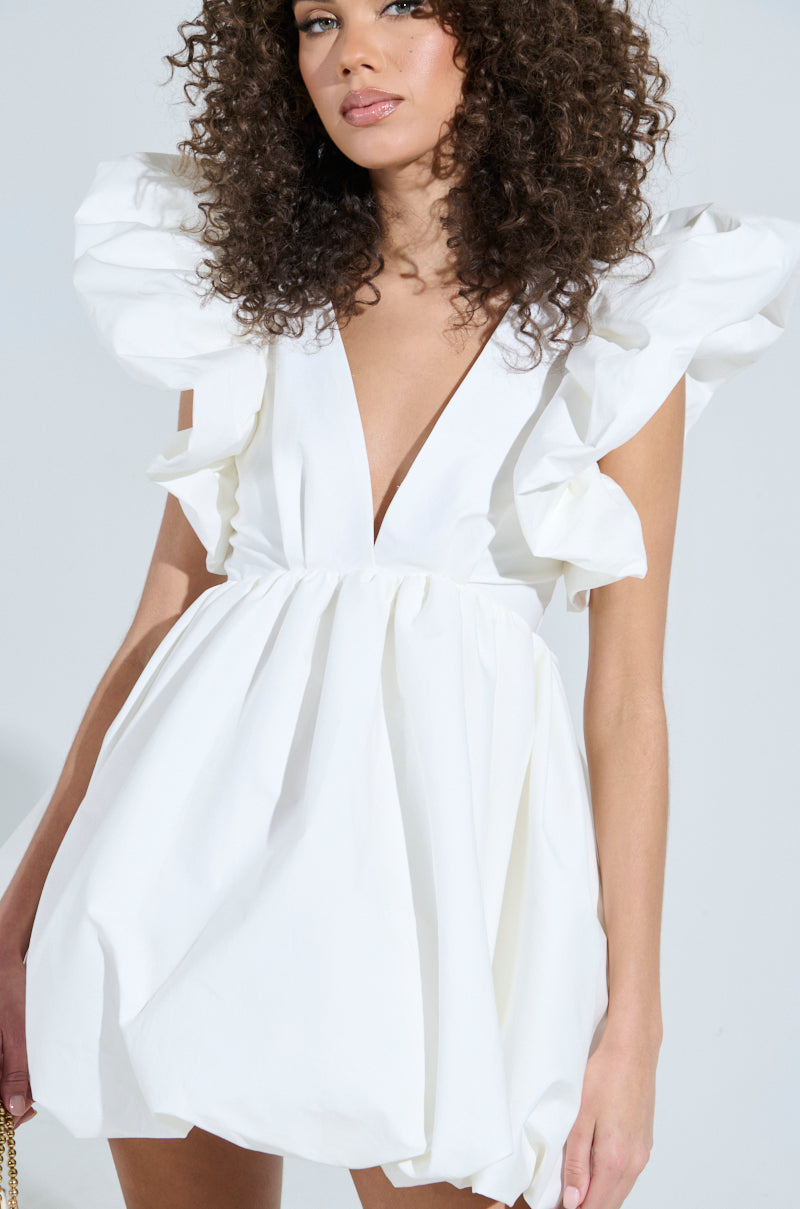 Wearing the MAIN CHARACTER ENERGY MINI DRESS IN WHITE, a woman with curly hair exudes confidence with its large ruffled shoulders and deep V-neckline. She complements her look by holding a small gold chain purse against a plain, light background. 7