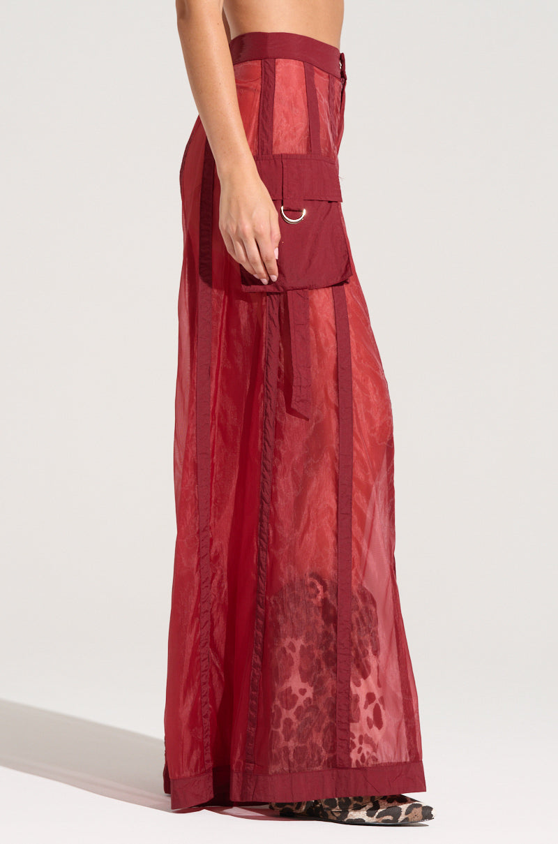 A model is showcasing the CELINE WIDE LEG TROUSER, a high-waisted red sheer pant with large vertical cargo-style pockets and leopard print lining. This floor-length design features intricate stitching details and a D-ring accent on the pocket flap, offering glimpses of the lower legs and feet through the sheer fabric.