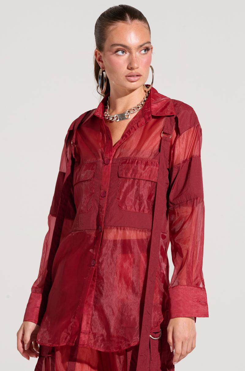 A model wearing the CELINE BUTTON DOWN SHIRT, a sheer, lightweight red blouse featuring utility-style breast pockets, paired with matching red pants. The outfit boasts textured accents and a loose fit. The model has straight hair pulled back into a ponytail and is accessorized with large hoop earrings and a chunky necklace.