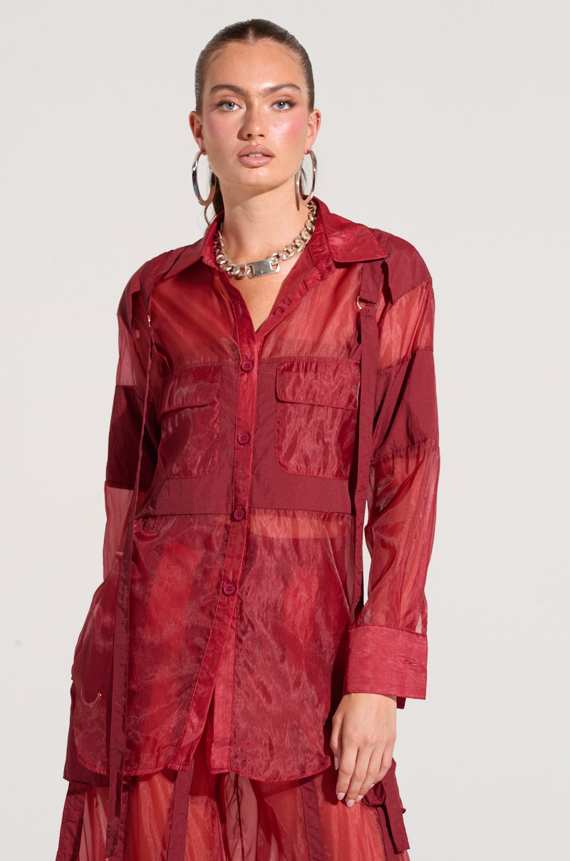 A person with shoulder-length hair is wearing a CELINE BUTTON DOWN SHIRT, which is a sheer lightweight blouse featuring long sleeves and utility-style breast pockets. They are accessorized with large hoop earrings and a chunky chain necklace. The background is plain and light-colored.