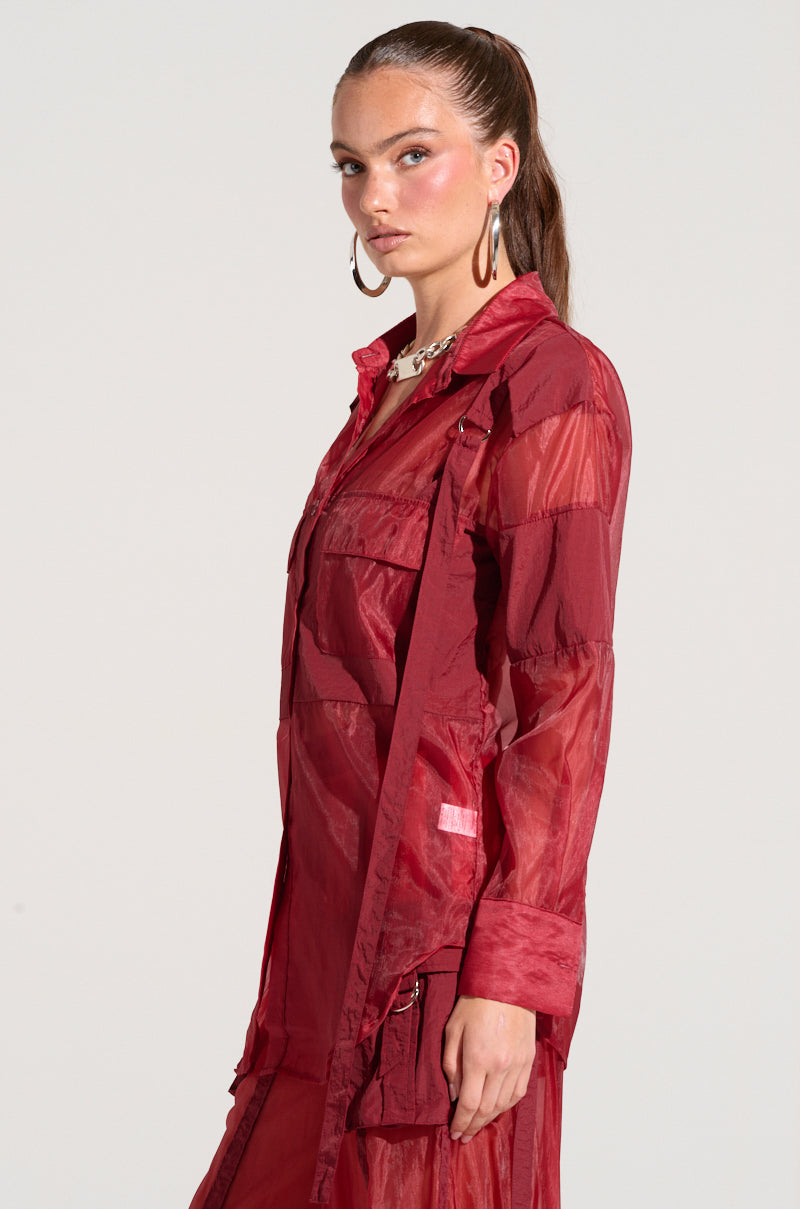 A person with long brown hair tied back in a ponytail wears the CELINE BUTTON DOWN SHIRT, a sheer, lightweight blouse featuring utility-style breast pockets. They stand in profile against a plain background, gazing towards the camera with a neutral expression. They accessorize with hoop earrings and a chunky necklace.