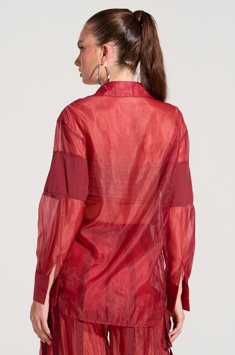 A person with long, brown hair tied in a ponytail is seen from the back, wearing a CELINE BUTTON DOWN SHIRT featuring sheer, lightweight fabric and solid red stripes on the sleeves. They have large hoop earrings and are standing against a plain background.