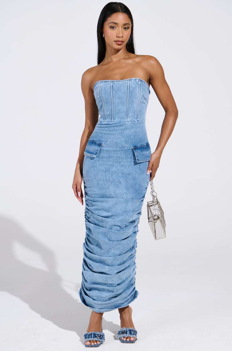 A woman models the ASH Strapless Maxi Dress in Denim, detailed with ruching at the bottom and hip pockets. She carries a small transparent handbag and wears matching denim sandals, set against a plain white background.