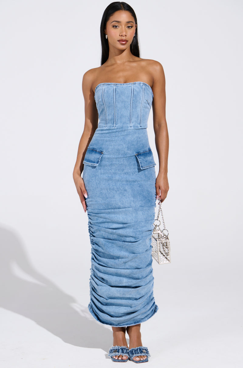 A woman wears the ASH STRAPLESS MAXI DRESS IN DENIM, featuring a ruched skirt and two front pockets. She holds a small purse and pairs it with embellished open-toed heels, standing against a plain white background.