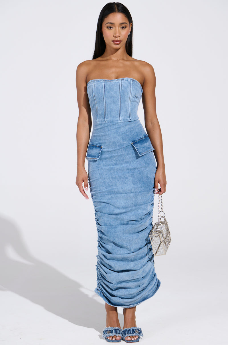 A woman poses in the ASH Strapless Maxi Dress in denim with ruched sides and two pockets, holding a small textured handbag and wearing matching denim shoes, set against a plain white background.