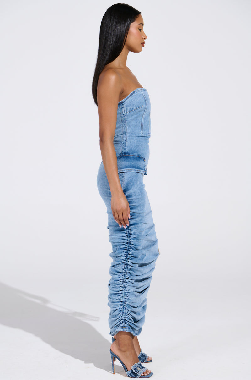 A woman in the ASH STRAPLESS MAXI DRESS IN DENIM stands facing left against a plain background. The dress features ruched leg detailing and is paired with high-heeled shoes. Her long, straight hair cascades down, highlighting the chic ensemble.