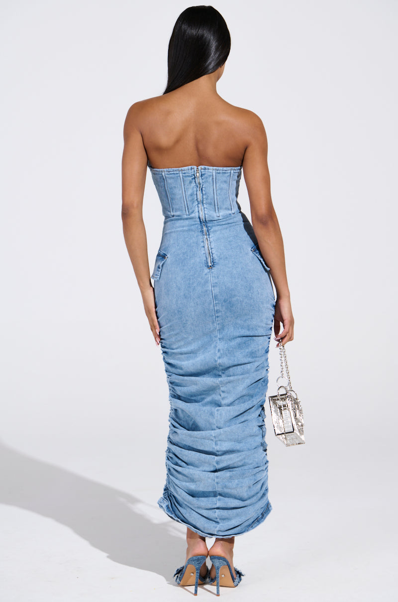 Rear view of a person wearing the ASH STRAPLESS MAXI DRESS IN DENIM, featuring a fitted bodice and gathered skirt with zipper and seam details. They hold a silver handbag, and long, straight hair cascades down their back.