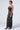 A person stands on a light gray background wearing the ASH Strapless Cargo Maxi Dress in black PU, an elegant, form-fitting garment that is gathered and ruched along the sides. Their hair is pulled back, and they accessorize with hoop earrings and pointed black shoes.