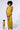 A woman stands against a plain backdrop wearing the ASH STRAPLESS CARGO MAXI DRESS, a mustard-yellow bodycon fit with ruching on the skirt. She holds a small, textured purse in her right hand. In the lower left corner, a smaller inset image displays the same outfit from another angle.