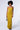 A woman with curly hair is wearing the ASH STRAPLESS CARGO MAXI DRESS, a bodycon fit, yellow strapless maxi dress featuring structured detailing and side flaps near the waist. She is holding a small, embellished handbag and standing against a plain white background.