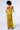 A woman stands with her back to the camera, highlighting the ASH Strapless Cargo Maxi Dress in a striking yellow hue. The dress, strapless and floor-length, features ruched detailing along the skirt that enhances its elegant design. With curly hair and strappy heeled sandals, she effortlessly showcases how the dress perfectly accentuates her figure against a plain white background.