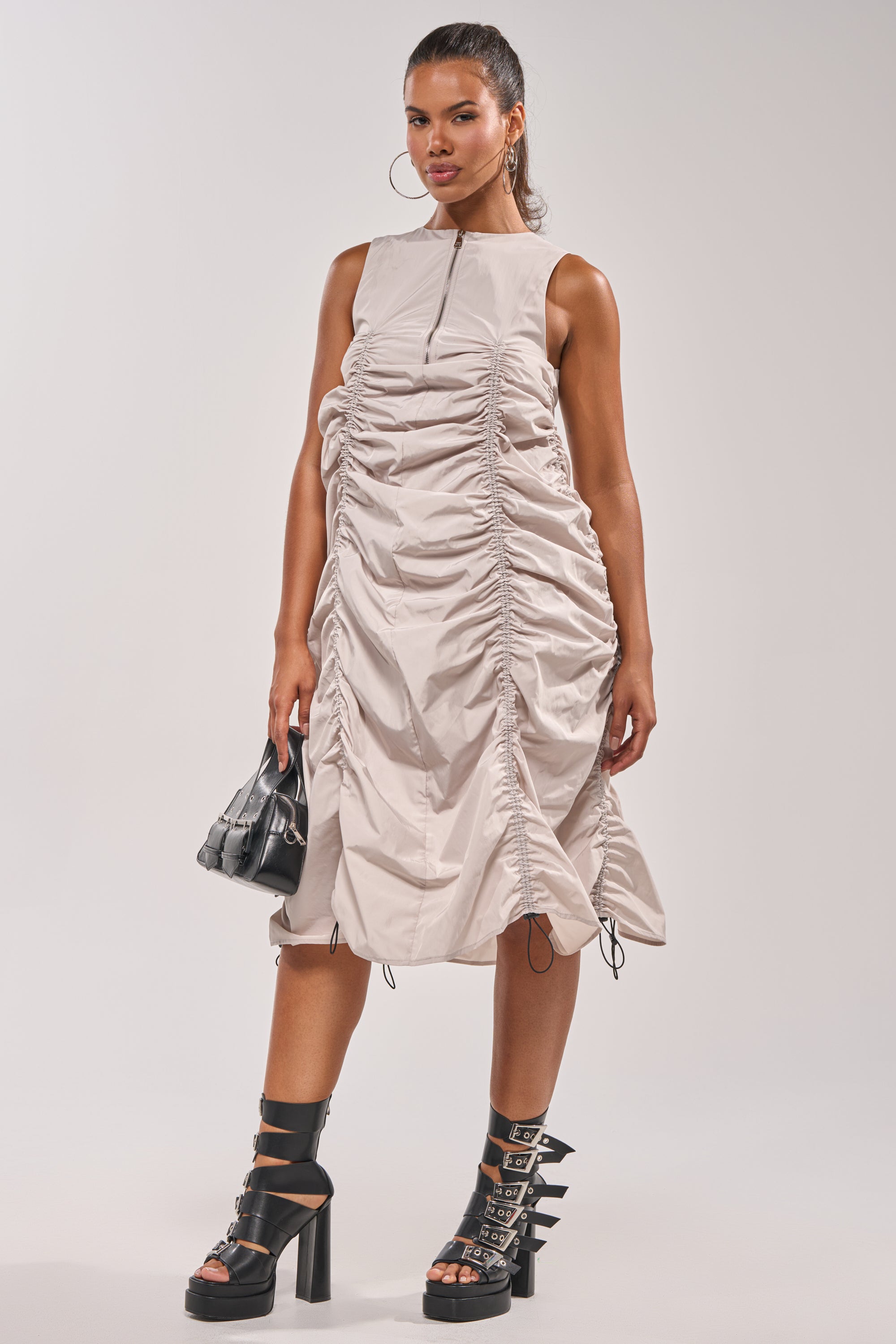 A woman stands confidently in the SORRY NOT SORRY NYLON CINCH MINI DRESS, styled with black platform heels, hoop earrings, and a small black handbag against a plain light background. 9